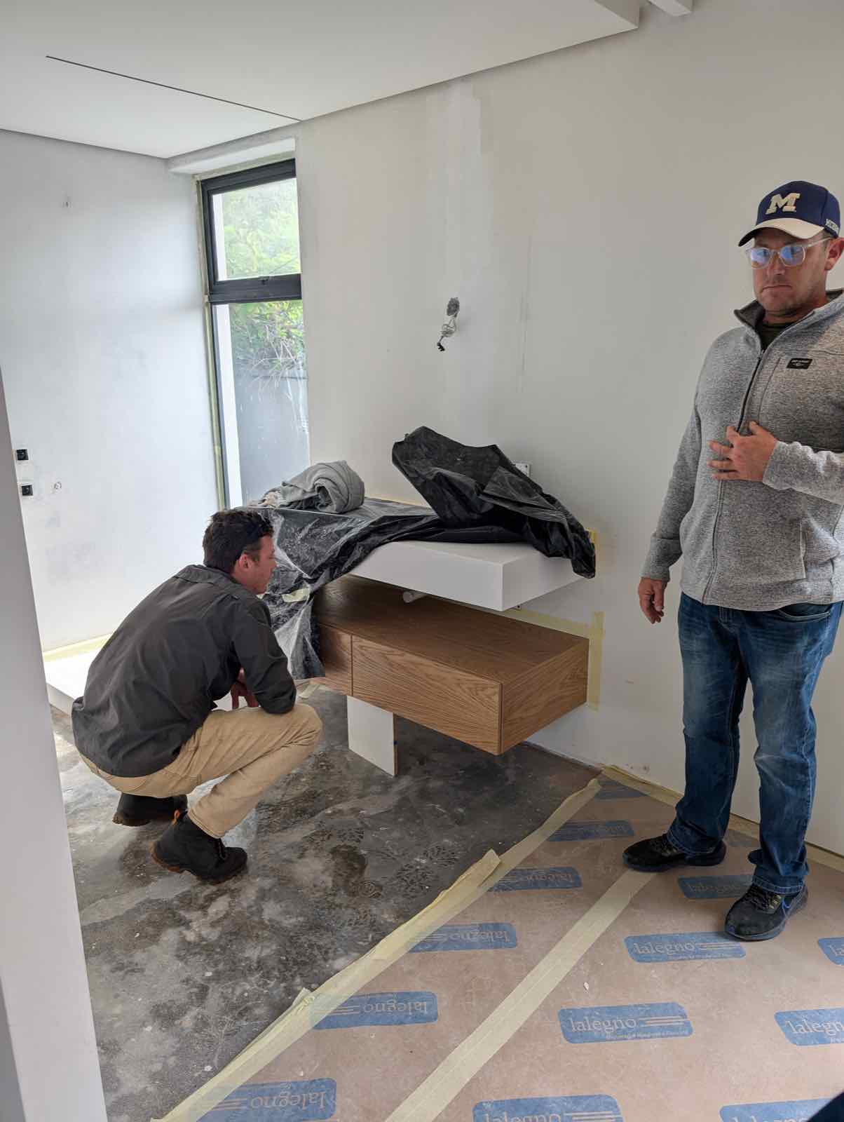 Worker crouched beside a newly installed floating oak bench in an unfinished room — protective paper and tape still on the floor