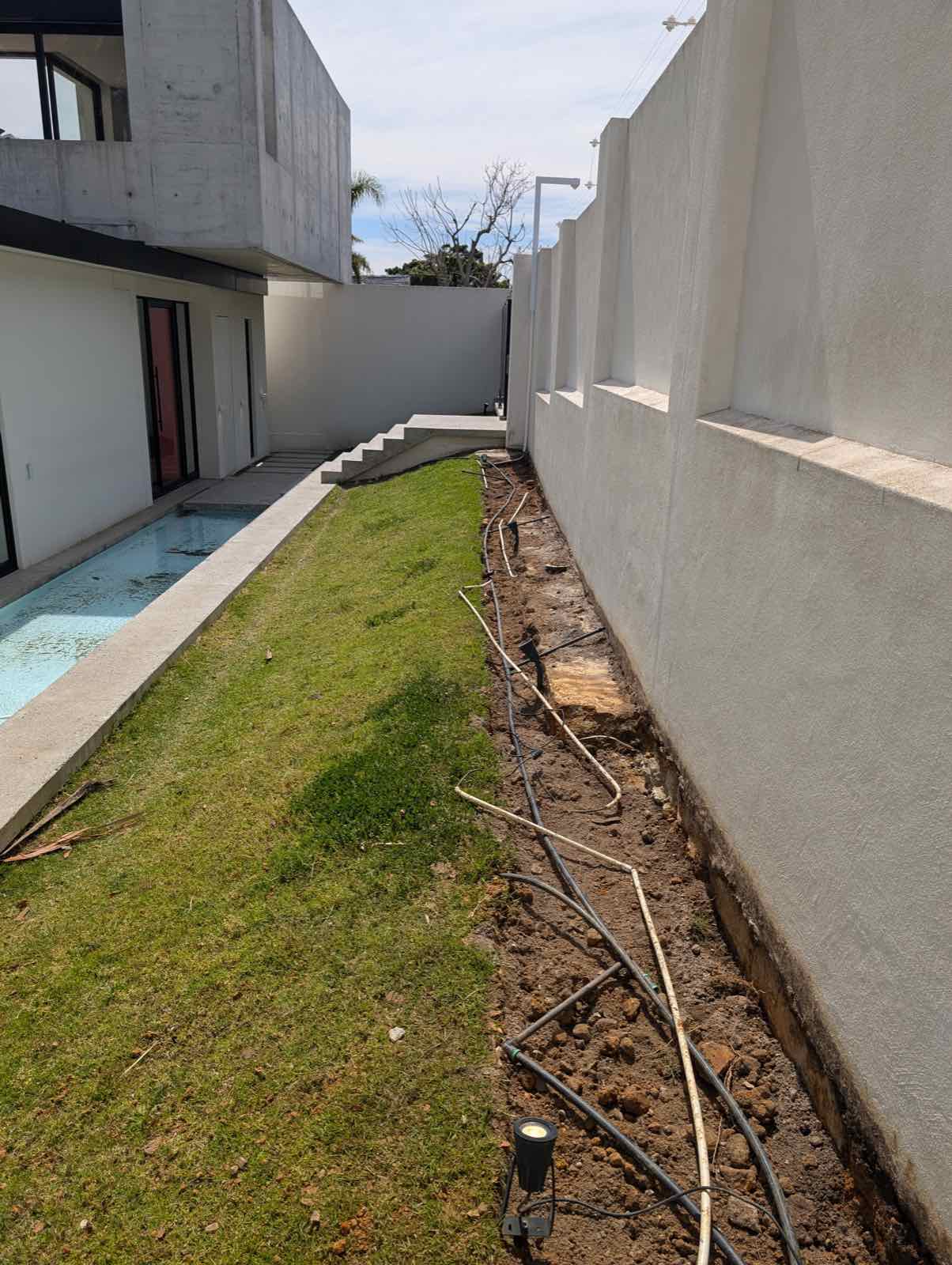 Raw irrigation trench cut through the grass along the bright white garden wall
